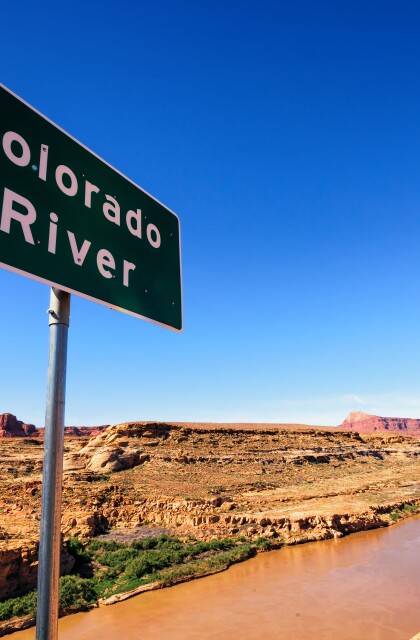 A sign identifying the Colorado River is located on a ridge overlooking the river surrounded by a desert landscape.