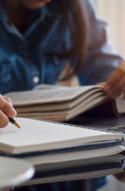 A student writes in a notepad while studying with a book and a computer. 