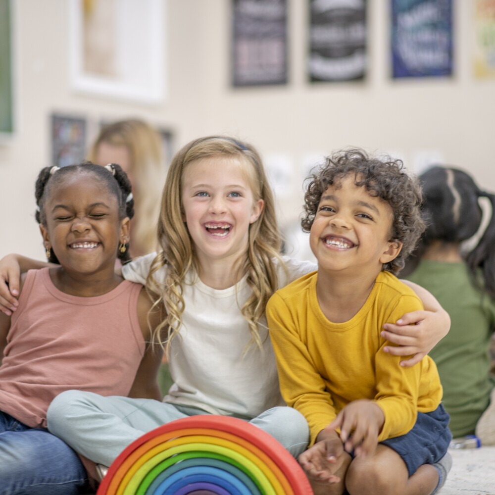 Three school students embrace in a classroom of children. 