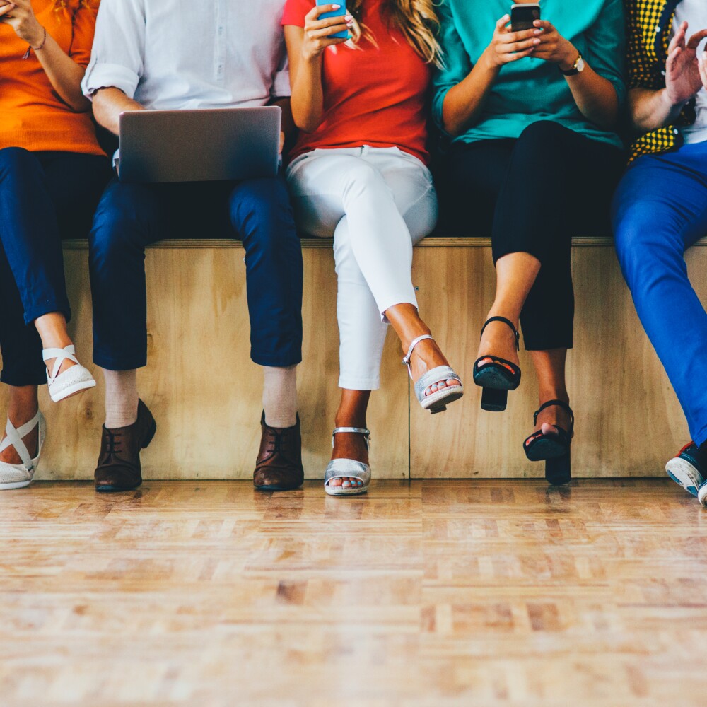 Students sit on a bench holding smart phones and desktop computers.