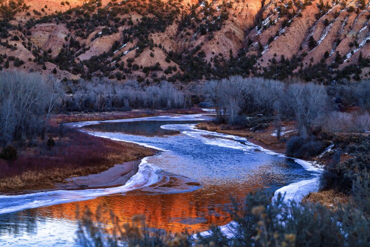 The Colorado River winds through a valley in winter, with ice along the river bank and mountains rising in the background. 