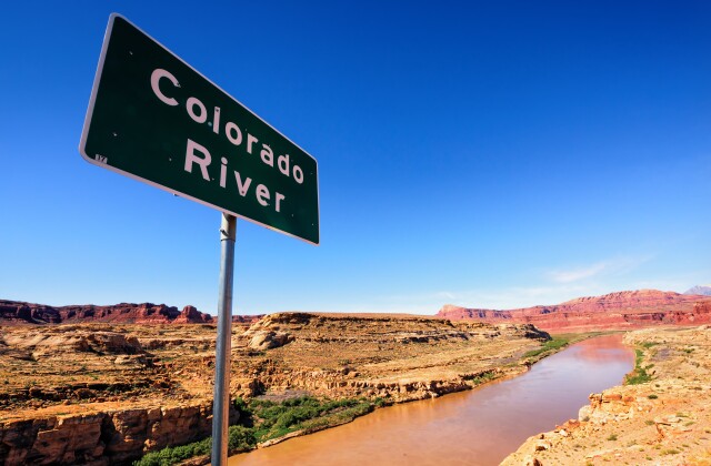 A sign identifying the Colorado River is located on a ridge overlooking the river surrounded by a desert landscape.