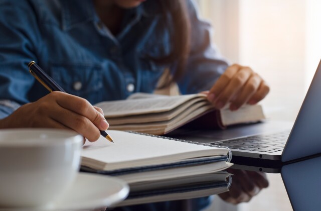 A student writes in a notepad while studying with a book and a computer. 