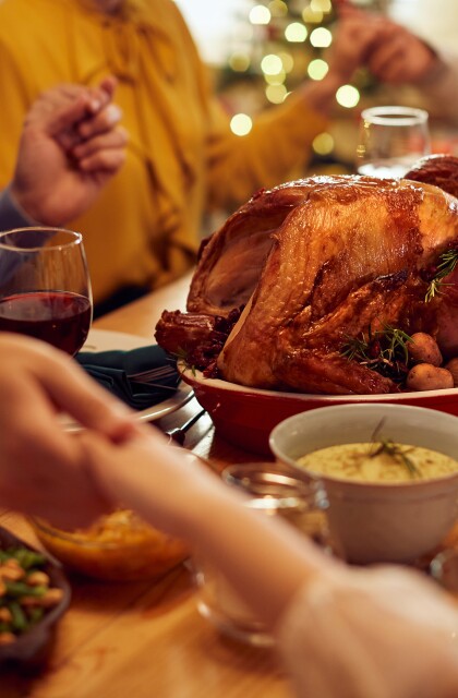 Family members hold hands around a Thanksgiving dinner table with turkey and side dishes. 