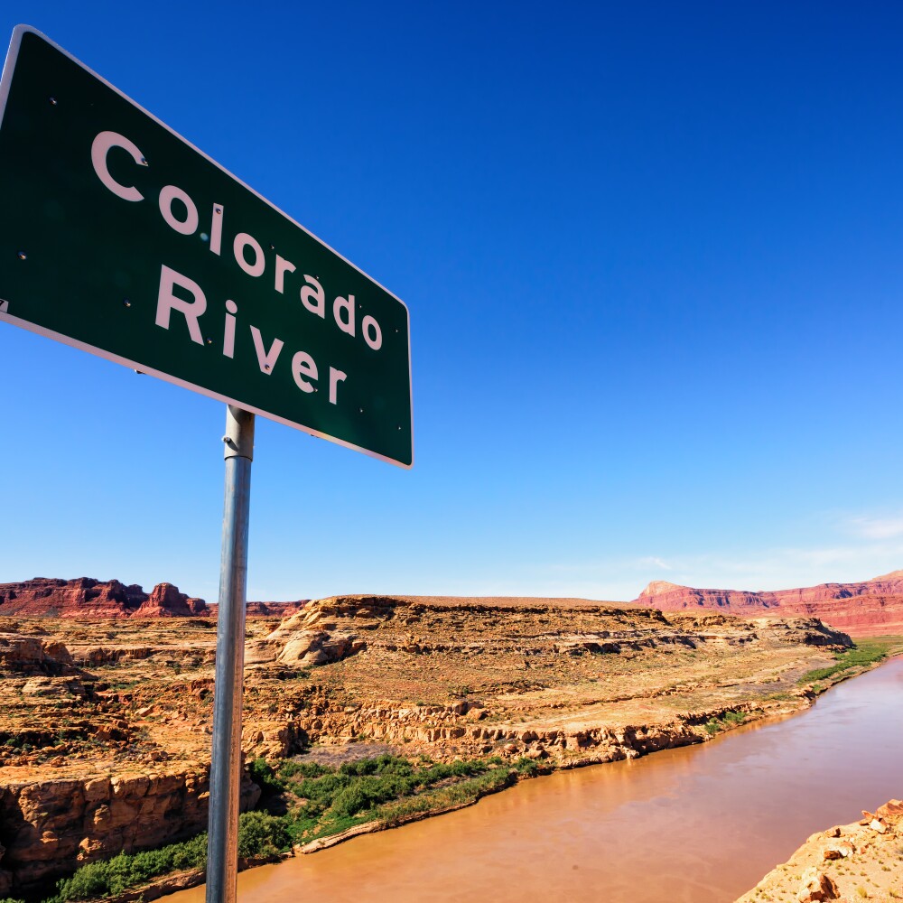 A sign identifying the Colorado River is located on a ridge overlooking the river surrounded by a desert landscape.