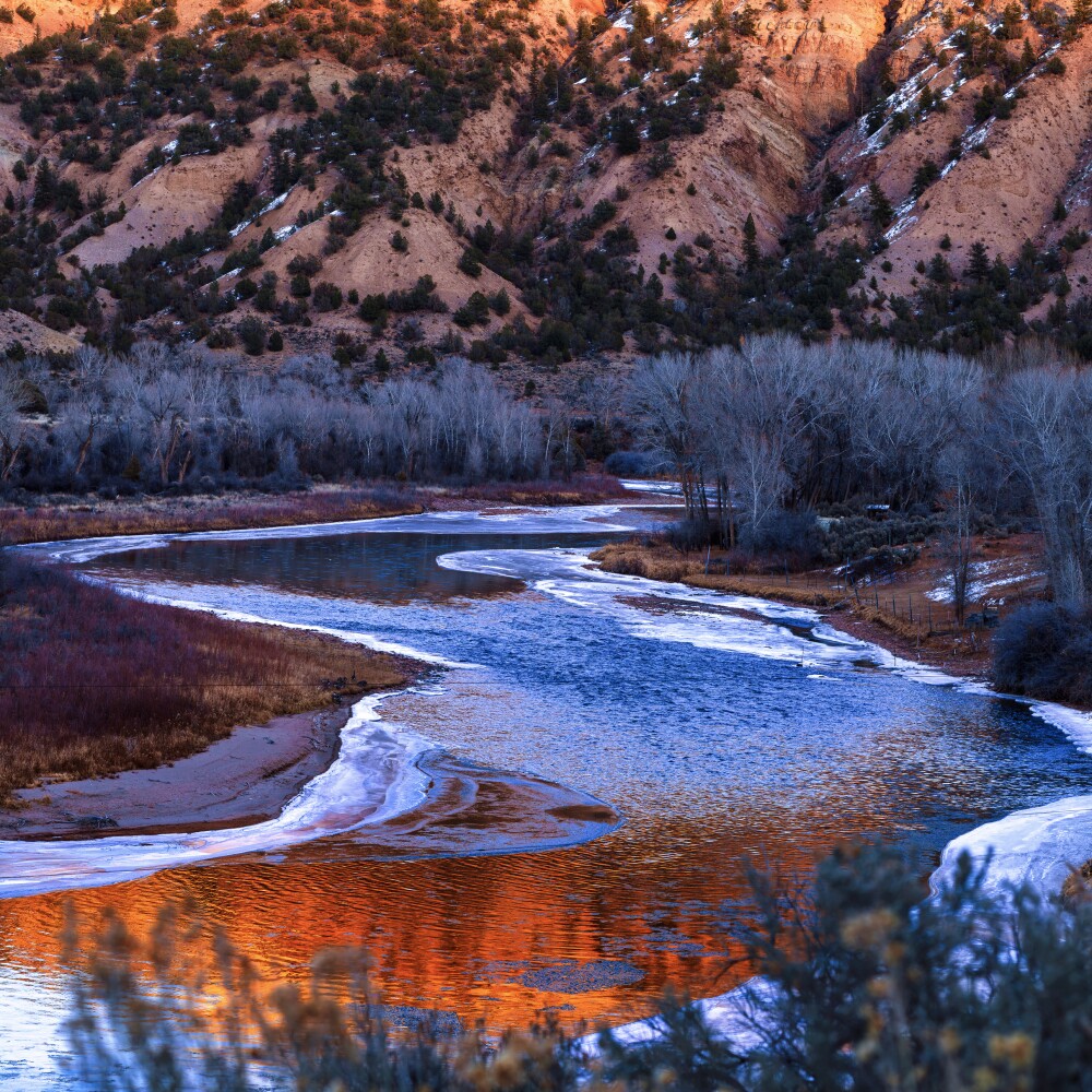 The Colorado River winds through a valley in winter, with ice along the river bank and mountains rising in the background. 