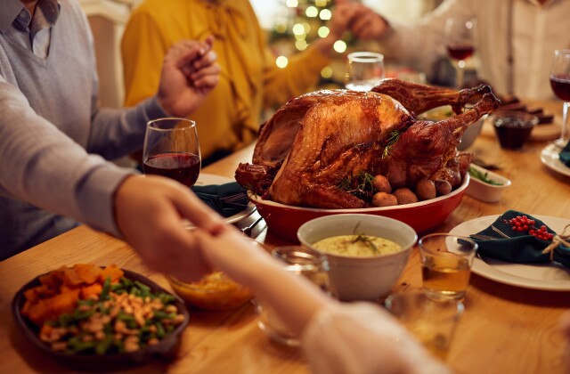 Family members hold hands around a Thanksgiving dinner table with turkey and side dishes. 
