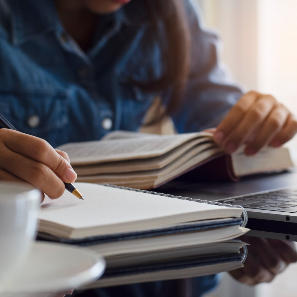 A student writes in a notepad while studying with a book and a computer. 