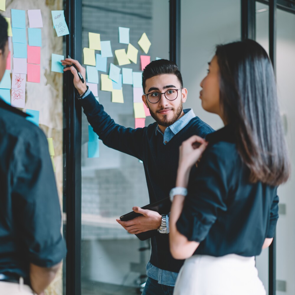 Three young adults talk beside a glass wall lined with sticky notes.